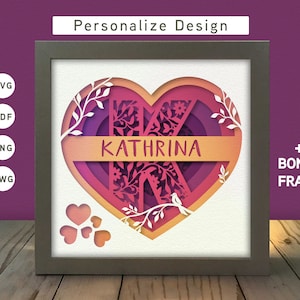 May include: A personalized heart-shaped frame with the name "KATHRINA" in a white banner. The heart is decorated with intricate floral designs and a bird. The frame is in a light brown wood finish.