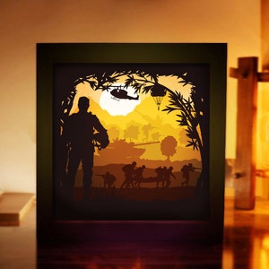 May include: A paper cut-out art piece depicting a soldier standing in a jungle scene. The soldier is silhouetted against a yellow and orange sunset. There are other soldiers in the background, as well as a helicopter and a hot air balloon.