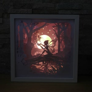 May include: A white framed paper light sculpture of a fairy with wings sitting on a branch in a forest with a moon and stars in the background.