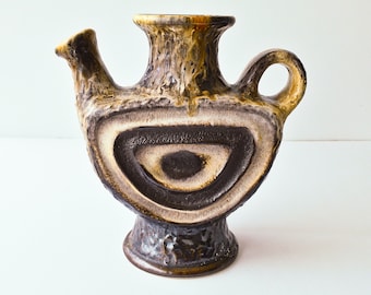 May include: A ceramic pitcher with a unique design. The pitcher features a spout, handle, and a wide opening. The body is decorated with concentric circles in shades of brown, beige, and black. The base and top have a textured, dripped glaze.