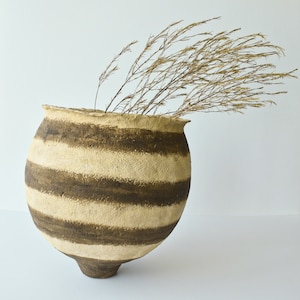 Puede incluir: Un jarrón de cerámica redondo hecho a mano, con una abertura ancha y un cuerpo bulboso. El jarrón presenta bandas horizontales alternas de beige y marrón oscuro. Hierbas secas de color beige están dispuestas dentro del jarrón, creando una estética natural y terrosa.