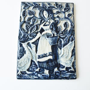 May include: A rectangular ceramic wall plaque with a relief design in shades of blue and white. The artwork depicts a woman in traditional attire with two geese, surrounded by foliage. The piece has a rustic, handcrafted appearance.