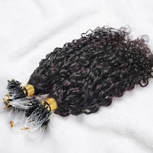 Könnte beinhalten: Nahaufnahme von dunklen, lockigen Haarverlängerungen. Die Extensions sind gebündelt und mit goldfarbenen Bändern befestigt. Das Haar hat eine tiefschwarze Farbe und ein enges, natürliches Lockenmuster. Der Hintergrund ist ein weicher, weißer Stoff.