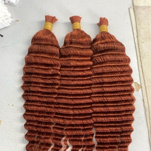 May include: Three bundles of copper-coloured human hair extensions in a wavy texture. The hair is tied together with a brown elastic band.