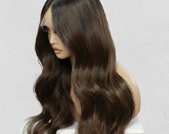 Può includere: Una lunga parrucca castana ondulata, acconciata su una testa di manichino. La parrucca presenta radici scure con lunghezze marroni più chiare, creando un naturale effetto ombré. I capelli sono acconciati con onde morbide e riga laterale.