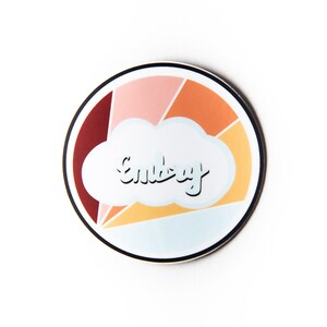 May include: A round sticker with a colorful geometric design in shades of red, orange, yellow, and pink. The design resembles a sunset with a white cloud in the center. The word "Embrace" is written in black cursive inside the cloud.