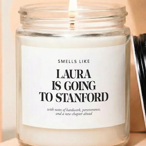 May include: A lit candle in a clear glass jar with a black lid. The label reads "SMELLS LIKE LAURA IS GOING TO STANFORD" with additional text. The candle sits on a peach-colored geometric display.