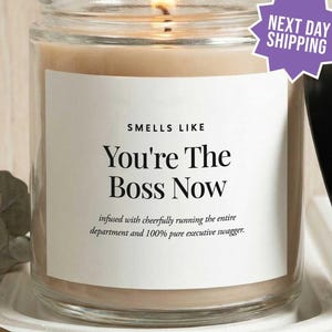 May include: A clear glass jar candle with a lit wick and a white label featuring the black text: SMELLS LIKE You're The Boss Now, infused with cheerfully running the entire department and 100% pure executive swagger. A black lid sits beside the jar.