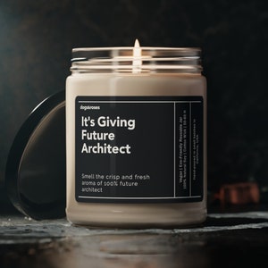 May include: A beige candle in a glass jar with a black label that reads "It's Giving Future Architect". The label also includes the text "Smell the crisp and fresh aroma of 100% future architect". The candle is lit and the flame is visible.