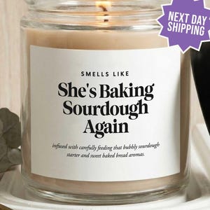 May include: A clear glass candle jar with a lit wick and a beige wax fill. The white label features black text reading: SMELLS LIKE She's Baking Sourdough Again, infused with carefully feeding that bubbly sourdough starter and sweet baked bread aromas.