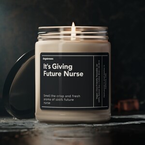 May include: A beige candle in a glass jar with a black label that reads "It's Giving Future Nurse" and "Smell the crisp and fresh aroma of 100% future nurse". The candle is lit and the flame is visible.