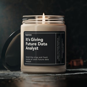 May include: A beige candle with a black label that reads "It's Giving Future Data Analyst". The label also includes the text "Smell the crisp and fresh aroma of 100% future data analyst". The candle is lit and the flame is visible.