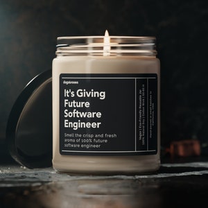 May include: A beige candle in a glass jar with a black label that reads "It's Giving Future Software Engineer" and "Smell the crisp and fresh aroma of 100% future software engineer". The candle is lit and the flame is visible.