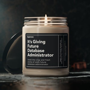 May include: A beige candle in a clear glass jar with a black label that reads "It's Giving Future Database Administrator" and "Smell the crisp and fresh aroma of 100% future database administrator". The candle is lit and the flame is visible.
