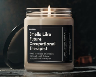 May include: A beige candle in a glass jar with a black label that reads "Smells Like Future Occupational Therapist". The label also says "Smell the crisp and fresh aroma of 100% future occupational therapist". The candle is lit and the flame is visible.