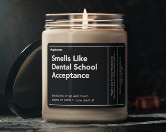 May include: A clear glass candle jar with a beige wax candle inside. The candle is lit and the jar has a black label that reads "Smells Like Dental School Acceptance" and "Smell the crisp and fresh aroma of 100% future dentist".