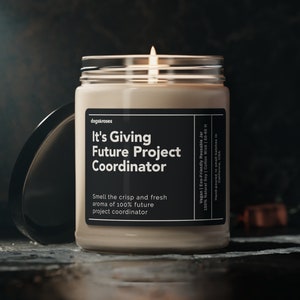 May include: A lit candle in a clear glass jar with a black lid. The label reads "It's Giving Future Project Coordinator" with the text "Smell the crisp and fresh aroma of 100% future project coordinator."