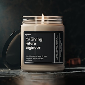 May include: A beige candle in a glass jar with a black label that reads "It's Giving Future Engineer". The label also includes the text "Smell the crisp and fresh aroma of 100% future engineer". The candle is lit and the flame is visible.