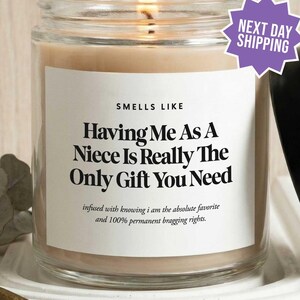 May include: A clear glass candle jar with a lit wick and a black lid. The white label features the text: SMELLS LIKE Having Me As A Niece Is Really The Only Gift You Need, infused with knowing i am the absolute favorite and 100% permanent bragging rights.