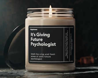 Puede incluir: Una vela beige con una etiqueta negra que dice "It's Giving Future Psychologist" y "Smell the crisp and fresh aroma of 100% future psychologist". La vela está encendida y la llama es visible. La vela está sobre una superficie oscura.