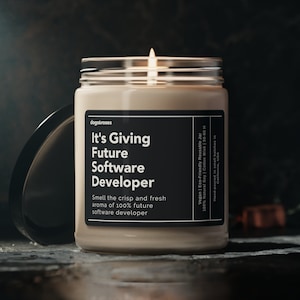 May include: A beige candle in a glass jar with a black label that reads "It's Giving Future Software Developer" and "Smell the crisp and fresh aroma of 100% future software developer". The candle is lit and the flame is visible.