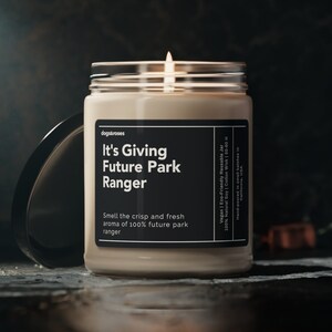 May include: A beige candle in a glass jar with a black label that reads "It's Giving Future Park Ranger" and "Smell the crisp and fresh aroma of 100% future park ranger". The candle is lit and the flame is visible.
