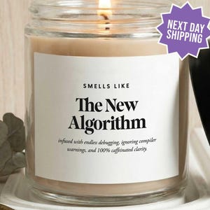 May include: A clear glass candle jar with a lit wick and a white label that reads: SMELLS LIKE The New Algorithm, infused with endless debugging, ignoring compiler warnings, and 100% caffeinated clarity. The candle has a black lid and beige wax.