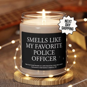 May include: A brown glass candle with a black label that reads "Smells Like My Favorite Police Officer". The candle is lit and sitting on a wooden coaster.