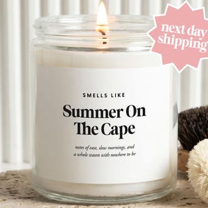 May include: A clear glass jar candle with a white label featuring the text "SMELLS LIKE Summer On The Cape" and "notes of ease, slow mornings, and a whole season with nowhere to be". The candle has a lit wick and a pink star-shaped sticker.