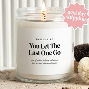 May include: A clear glass candle jar with a white label featuring the black text "SMELLS LIKE You Let The Last One Go" and "notes of stillness, disbelief, and a house that has never once been this quiet." The candle has a lit wick.
