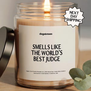 May include: A lit candle in a glass jar with a black lid. The label reads "SMELLS LIKE THE WORLD'S BEST JUDGE" with the brand name "dogs&roses". A "NEXT DAY SHIPPING" sticker is visible. The candle is on a wooden surface.
