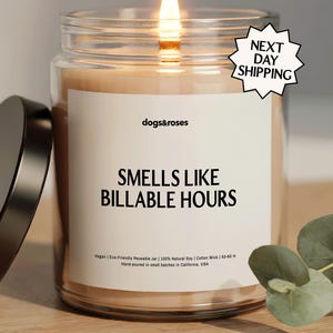 May include: A lit candle in a clear glass jar with a black lid. The label reads "dogs&roses" and "SMELLS LIKE BILLABLE HOURS." The candle is on a wooden surface. A sprig of eucalyptus is in the background.