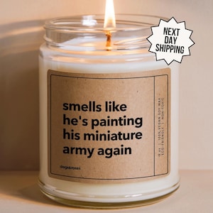 May include: A white candle with a brown label that reads "smells like he's painting his miniature army again". The candle is in a clear glass jar with a gold lid. The label also says "9 oz | 100% Vegan Soy Wax | Eco-Friendly | Non-Toxic" and "dogs&roses".