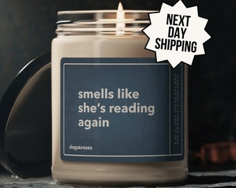 May include: A soy wax candle with a blue label that reads "smells like she's reading again" and "dogs&roses". The candle is lit and has a white wax exterior. The label also includes the text "9oz 100% VEGAN SOY WAX ECO-FRIENDLY NON-TOXIC".