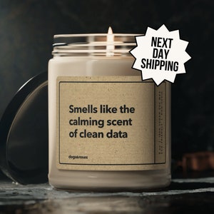 May include: A beige candle with a brown label that reads "Smells like the calming scent of clean data" and "dogs&roses". The candle is lit and has a white wick. The label also includes the text "90-100% VEGAN SOY WAX ECO-FRIENDLY NON-TOXIC".