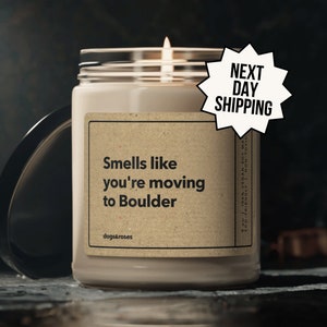 May include: A brown candle with a white label that reads "Smells like you're moving to Boulder" and "dogs&roses". The candle is lit and has a white lid. The label also says "9 oz 100% Vegan Soy Wax Eco-Friendly Non-Toxic".