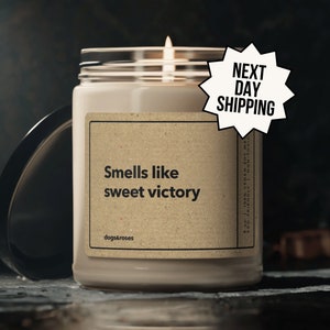 May include: A brown glass candle with a white label that reads "Smells like sweet victory" and "dogs&roses". The candle is lit and has a white flame. The candle is on a dark surface.