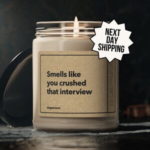May include: A brown soy wax candle with a white label that reads "Smells like you crushed that interview" and the brand name "dogs&roses". The candle is lit and has a white flame. The candle is on a dark surface with a black lid next to it. A white starburst sticker with the text "NEXT DAY SHIPPING" is on the right side of the image.