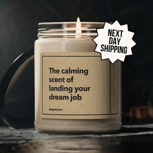 May include: A beige candle with a brown label that reads "The calming scent of landing your dream job" and "dogs&roses". The candle is lit and has a clear glass jar.