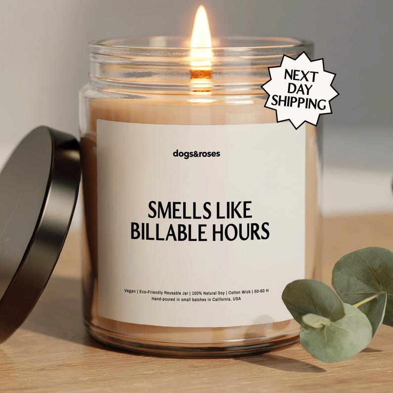 May include: A lit candle in a clear glass jar with a black lid. The label reads "dogs&roses" and "SMELLS LIKE BILLABLE HOURS." The candle is on a wooden surface. A sprig of eucalyptus is in the background.
