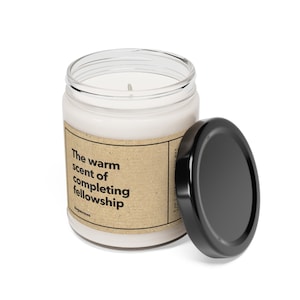 May include: A white candle in a glass jar with a brown paper label that reads "The warm scent of completing fellowship". The candle has a black lid.