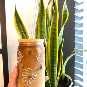 May include: A glass of iced coffee in a cup with a bamboo lid and a clear glass straw. The cup has a white outline of tropical leaves on it. The cup is being held by a hand.