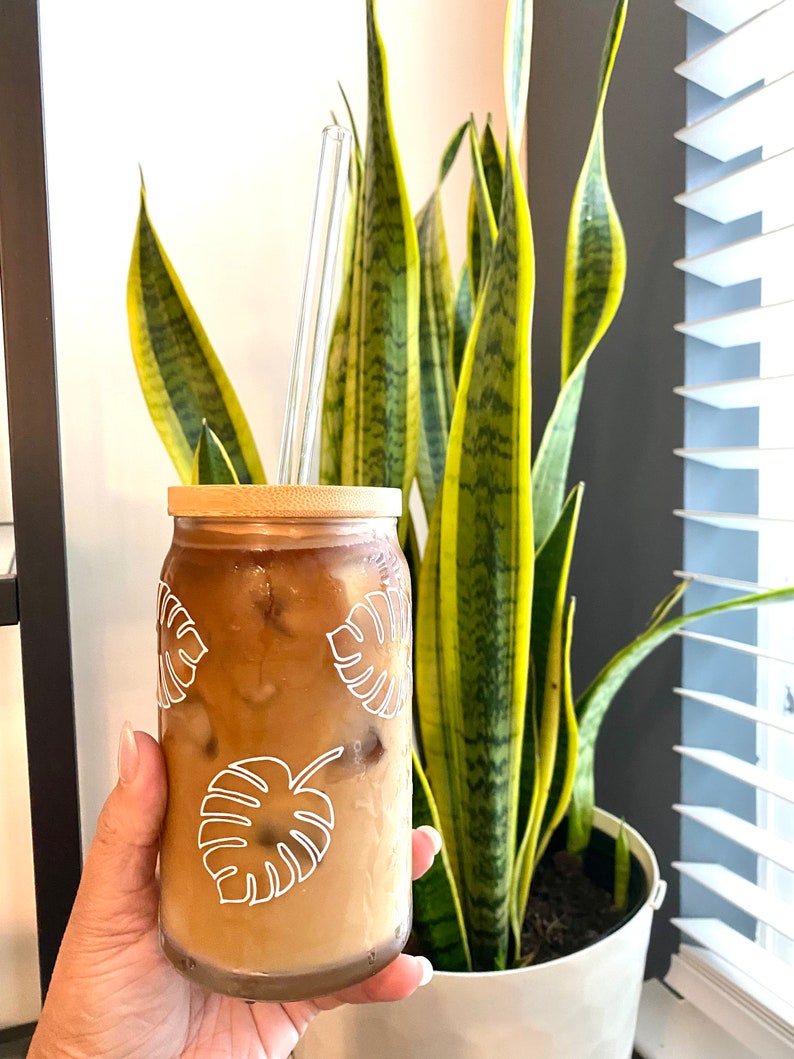 May include: A glass of iced coffee in a cup with a bamboo lid and a clear glass straw. The cup has a white outline of tropical leaves on it. The cup is being held by a hand.