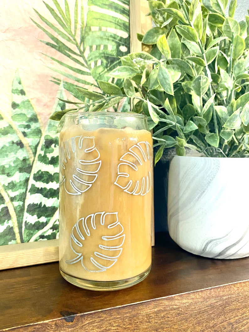 May include: A glass can containing a brown iced coffee drink. The can has a white outline of tropical leaves on the side.
