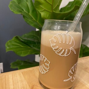 May include: A glass with a brown beverage and a clear drinking straw. The glass has a white design of tropical leaves on it.