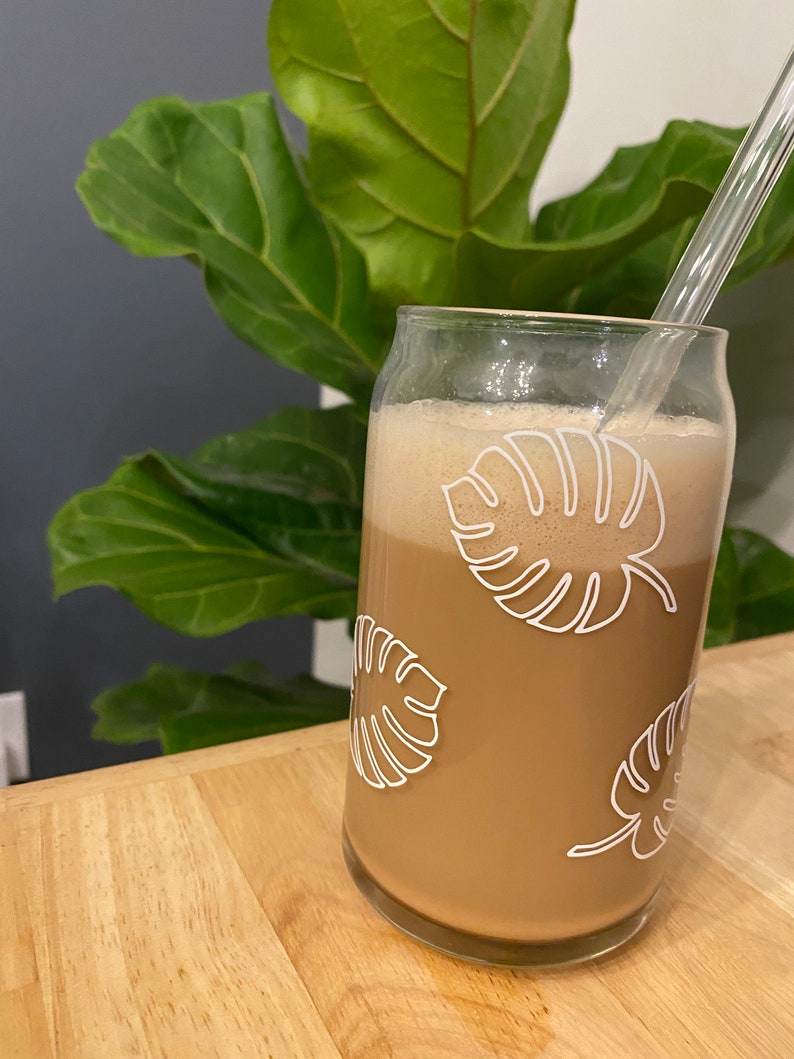 May include: A glass with a brown beverage and a clear drinking straw. The glass has a white design of tropical leaves on it.