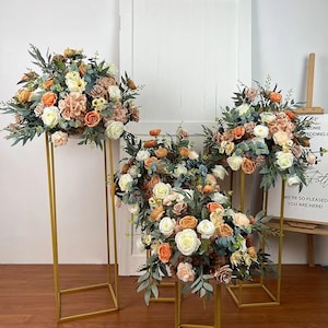 May include: Four gold metal stands with floral arrangements on top. The flowers are a mix of peach, white, and cream roses, eucalyptus, and other greenery. The stands are on a wooden floor in front of a white door.