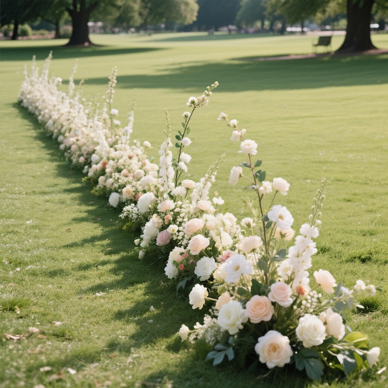 Puede incluir: Un arreglo floral largo y bajo de rosas blancas y rosas p&aacute;lidas, y otras flores, dispuesto sobre un c&eacute;sped verde. Las flores est&aacute;n dispuestas en l&iacute;nea, creando un camino o borde. El fondo presenta un campo de hierba y &aacute;rboles.