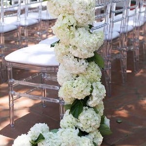 May include: Clear acrylic chairs with white cushions are adorned with a garland of white hydrangea flowers and green leaves. The floral display cascades down the chairs, creating a decorative arrangement for an event.