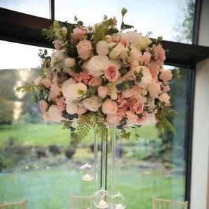 Könnte beinhalten: Eine hohe, klare Glasvase gefüllt mit einem großen Arrangement aus rosa und weißen Blumen. Die Blumen sind in einer kaskadierenden Form angeordnet, wobei die Stiele aus der Vase herausragen. Die Vase steht auf einem Tisch vor einem Fenster.